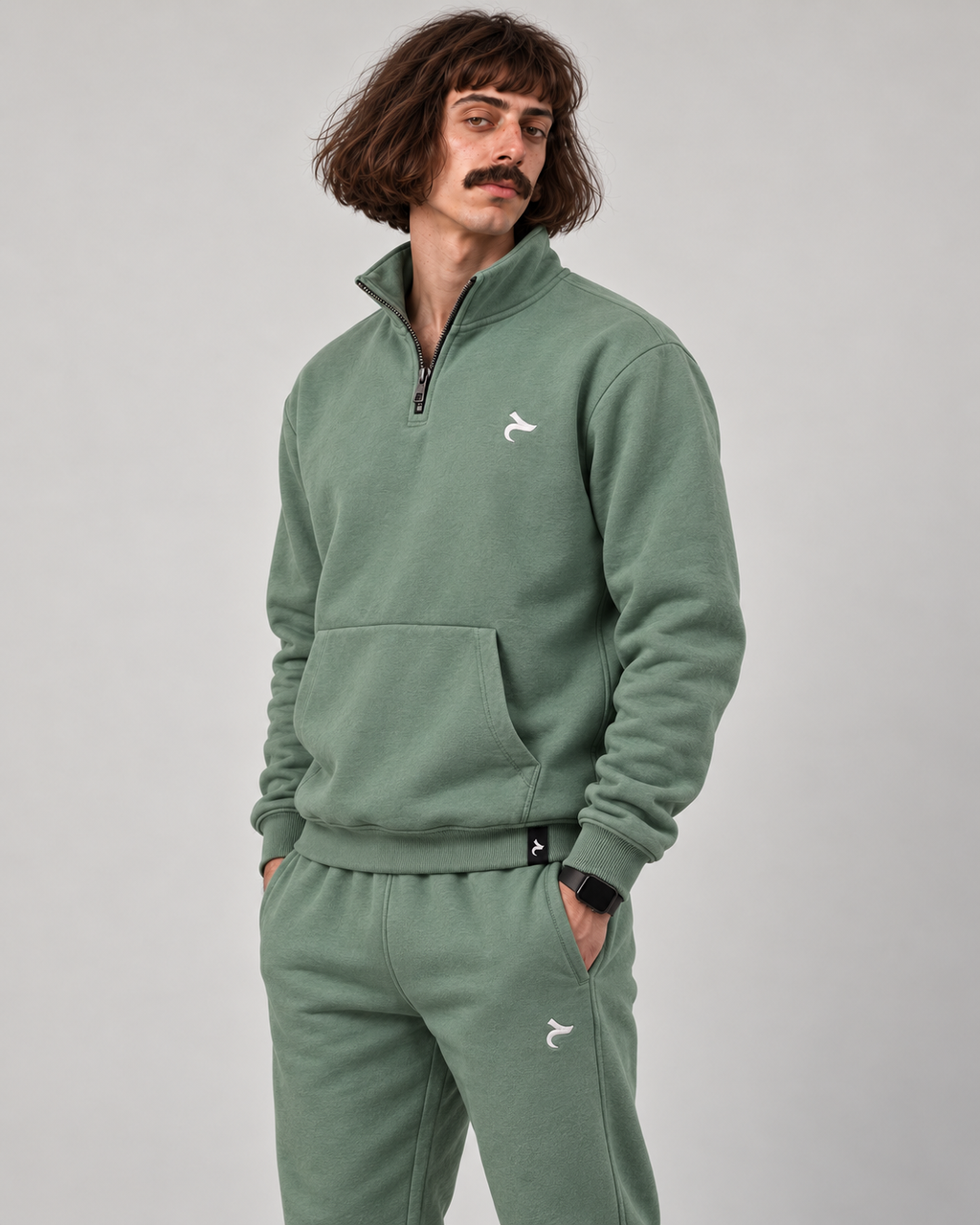A man wearing a sage green fleece tracksuit with a matching half-zip pullover and sweatpants, featuring a small white embroidered logo on the chest and thigh, set against a plain gray studio background.